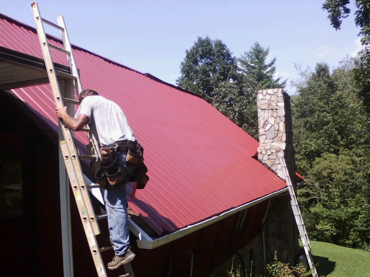 Roofer on a ladder beside a red metal roof and stone chimney — Soffit & Fascia Repair by Roofing Brothers in Labadie